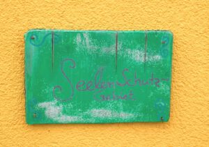Grüne Holztafel auf der gelben Hauswand der Praxis für Körpertherapie in Ettenheimweiler