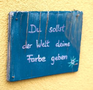 Blaues Holzschild mit dem Spruch "Du sollst der Welt deine Farbe geben" in der Körpertherapie Praxis in Ettenheimweiler